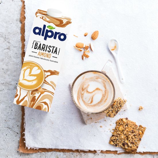 Alpro Barista Amandelmelk naast een kop koffie met latte art en gebroken amandelen. Pakket met marmerprint.