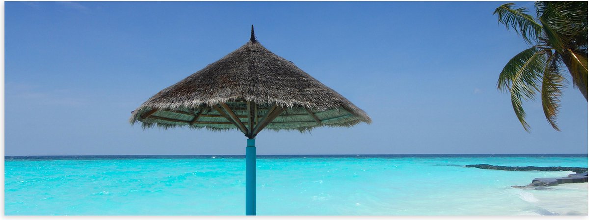 Poster Glanzend – Rieten Parasol op het Strand bij het Blauwe Water - 90x30 cm Foto op Pos