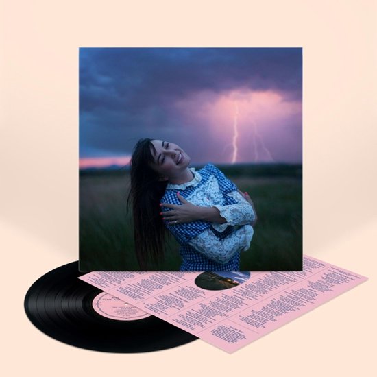 Een vrouw met lang bruin haar, gekleed in een blauw-wit geruite jurk met witte kanten details, omhelst zichzelf met gesloten ogen, terwijl bliksem in de verte de lucht verlicht.