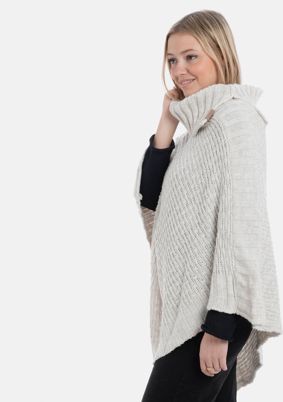 Lichtbeige gebreide poncho met hoge sjaalkraag, gedragen door een blonde vrouw, gefotografeerd vanaf de zijkant.