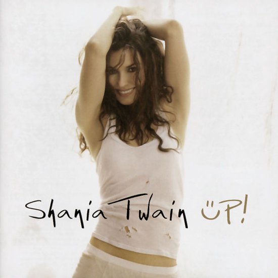 Shania Twain draagt een witte tanktop en lichtbruine broek, handen in haar haar, glimlachend.