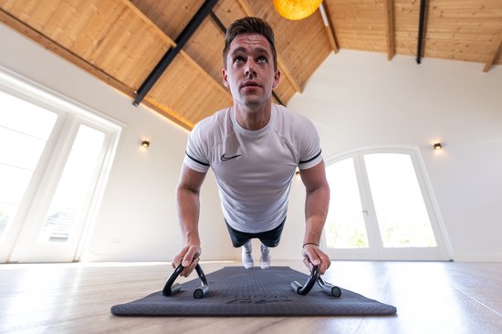 Een man in een wit shirt doet een push-up met zwarte opdruksteunen op een mat, in een kamer met houten plafond.