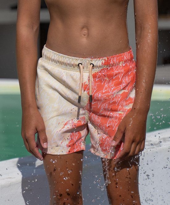 Jongen in zwembroek met half koraalrode, half gele print. Elastische tailleband met trekkoord.