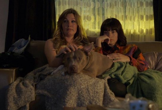 Twee vrouwen en hond op bank, kijkend naar tv. Vrouw met blond haar, met bloemenjurk.