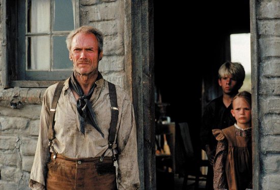 Clint Eastwood als "The Schofield Kid" met een zonnebloem op zijn schouder, staand voor een stenen muur, met een jongen en meisje op de achtergrond.