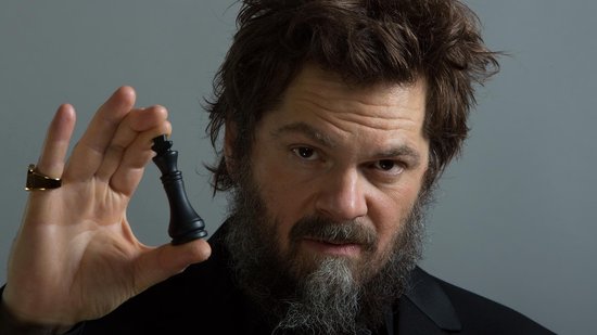 Een man met een baard en warrig haar houdt een zwarte koningsschaakstuk vast.