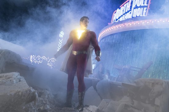 Shazam, in zijn rode pak met cape, staat tussen puin met op de achtergrond een verlichte kermisattractie.
