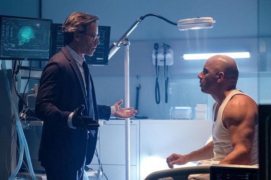 Man in pak die praat met Vin Diesel in witte tanktop in een laboratorium.