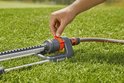 Een hand stelt een oranje knop in op een Gardena zwenksproeier op gras.