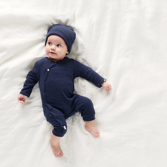 Baby in een donkerblauw boxpak en bijpassende muts, liggend op een wit laken.