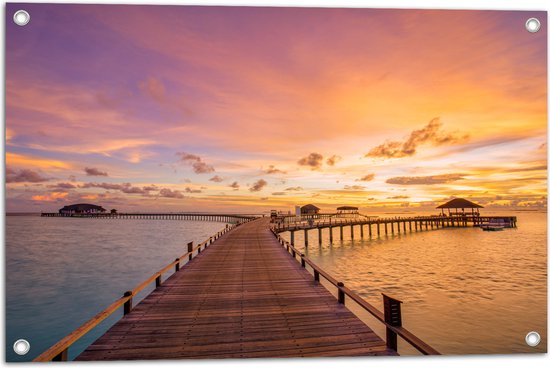Een houten pier strekt zich uit naar de oceaan, onder een oranje en paarse zonsondergang met gebouwen op palen.