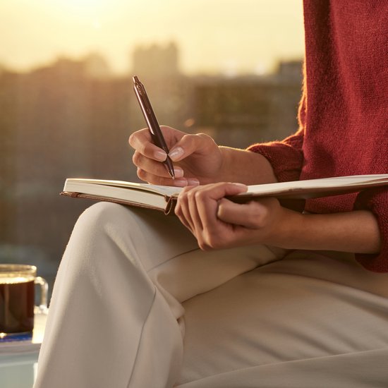 Femme portant un pull rouge écrit dans un carnet ouvert avec un stylo bordeaux. Coucher de soleil en arrière-plan.