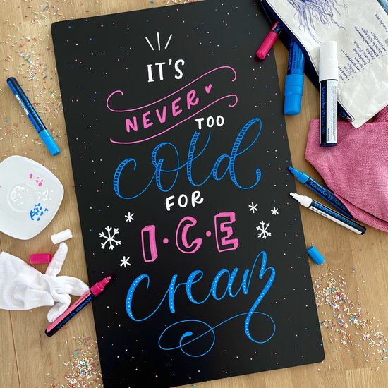 Tableau noir avec le texte "IT'S NEVER TOO COLD FOR ICE CREAM" en lettres blanches, roses et bleues. Autour du tableau se trouvent des vermicelles colorés, des lingettes et des marqueurs.
