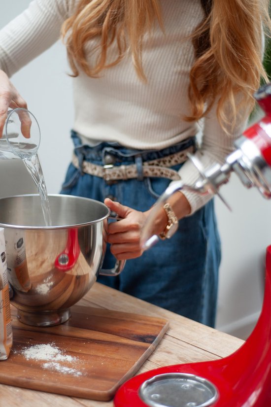 Vrouw voegt water toe aan een metalen mixerkom met meel, naast een rode keukenmachine.