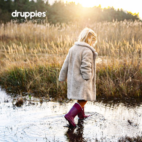 Meisje met blond haar in een lichte teddy jas, paarse regenlaarzen, loopt door de modder en het water in een rietveld.