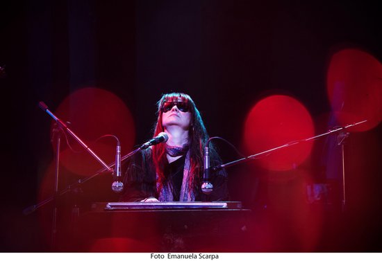 Een vrouw met lang haar en zonnebril speelt keyboard in een verlichte studio met rode lichten.