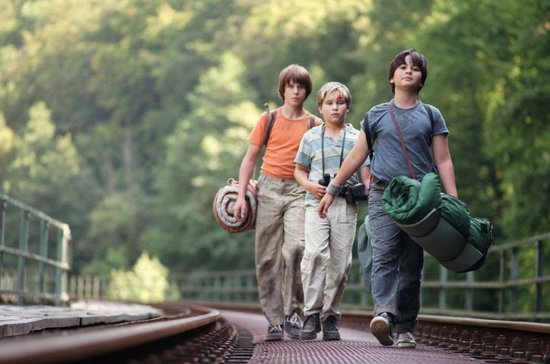 Drie jongens lopen over een spoorlijn met rugzakken en slaapzakken.