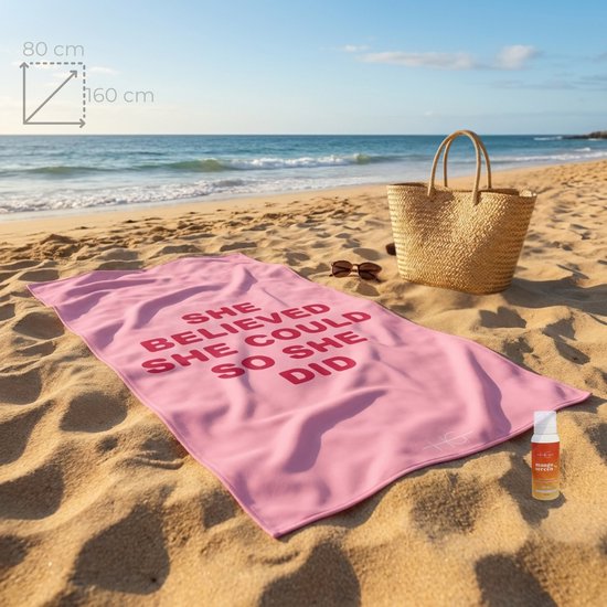 Roze microvezel handdoek met de tekst 'SHE BELIEVED SHE COULD SO SHE DID' op een zonnig strand. Een rieten tas, zonnebril en een product staan ernaast.