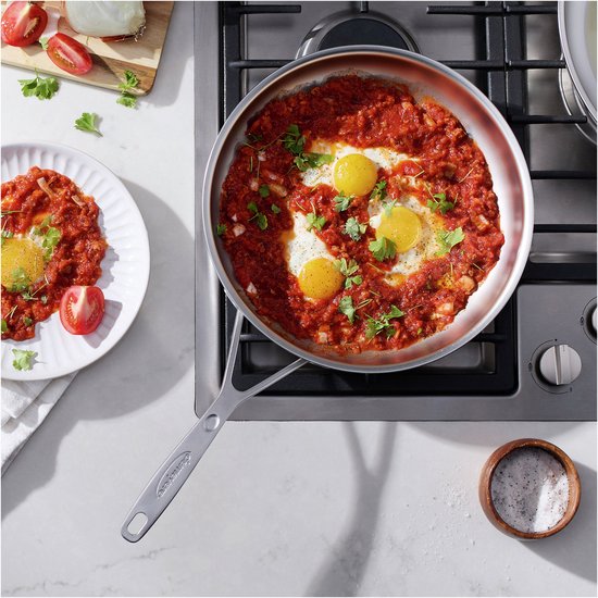 Roestvrijstalen koekenpan met shakshuka, witte bord met shakshuka en hand op het fornuis.
