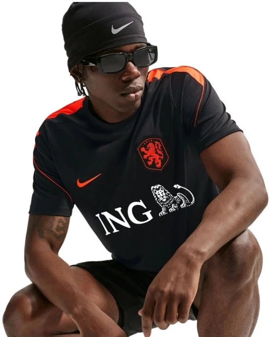 Model zittend in zwart sportshirt met oranje schouderdetails, Nederlandse leeuw en "ING" opdruk.