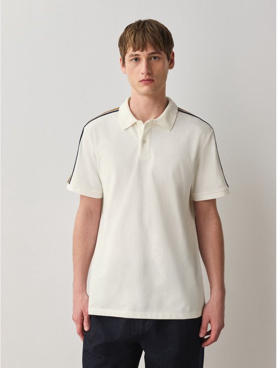Man met kort bruin haar draagt een crème poloshirt met strepen op de schouders en mouwen.