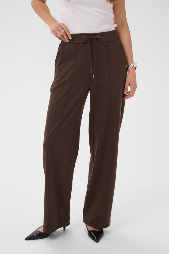 Model in een bruine broek met strikceintuur, witte tanktop en zwarte puntige schoenen.