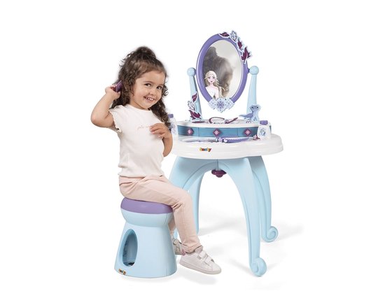 Een meisje met krullend haar zit op een blauw krukje en kamt haar haar bij een witte Frozen-kaptafel met spiegel.