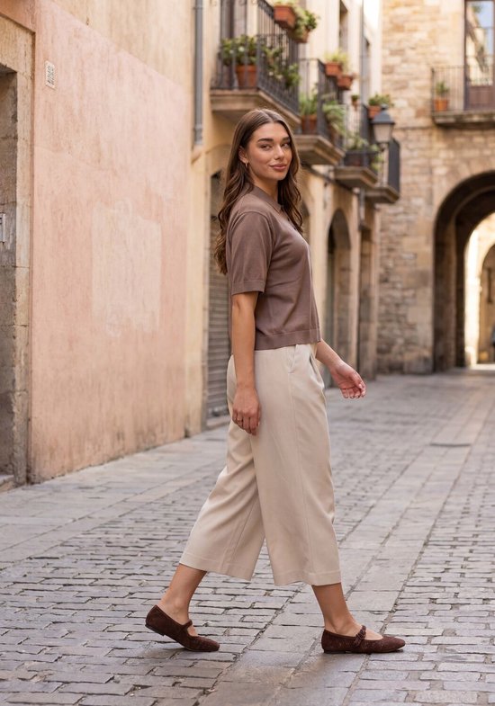 Model loopt in een beige culotte broek en bruin poloshirt op een straat met stenen.