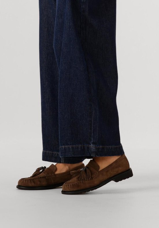 Close-up van de voeten van een model die donkerblauwe jeans draagt, met bruine suède loafers met kwastjes.