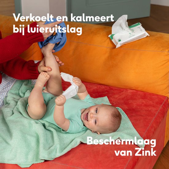 Baby lacht terwijl zijn luier wordt verschoond op een oranje bank. Naïf billendoekjes en crème zichtbaar.