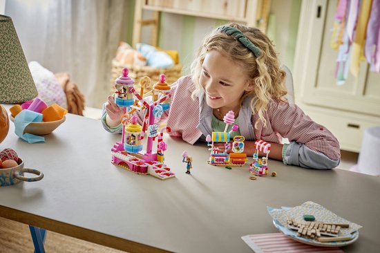 Een meisje met blond krullend haar, een groene haarband en een gestreepte blouse speelt met een LEGO Friends reuzenrad van snoepjes.