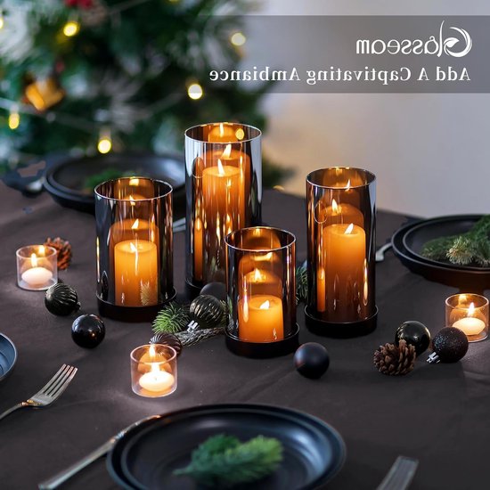 Een gedekte tafel met vier glazen kaarsen houders met grijze glazen kap, zwarte basis, oranje kaarsen erin, dennenappels en kerstballen.