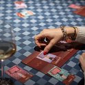 Een vrouw met gouden armband speelt een kaartspel aan een tafel met een blauw-wit geblokt tafelkleed. Ze legt een kaart op het speelbord.