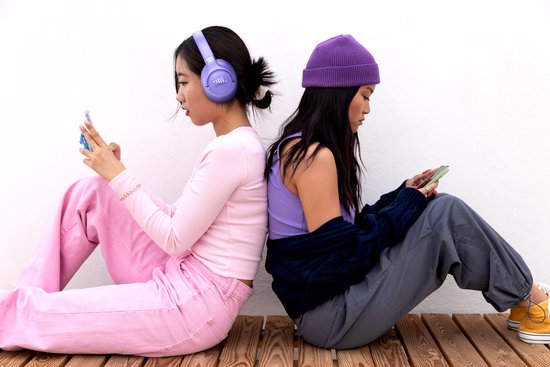 Twee vrouwen zitten rug aan rug. De linker draagt een paarse koptelefoon.