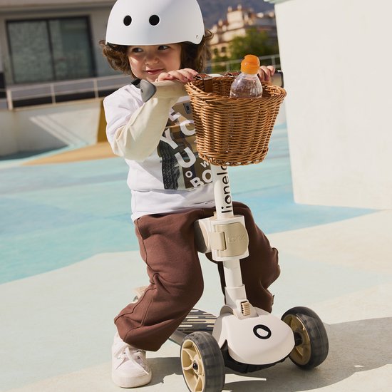 Jongen met witte helm en bruine joggingbroek op een roze driewielige step met rieten mandje.