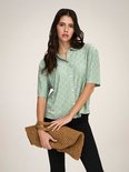 Model met een rieten tas in handen, in een lichtgroene blouse met broderie anglaise en korte mouwen.