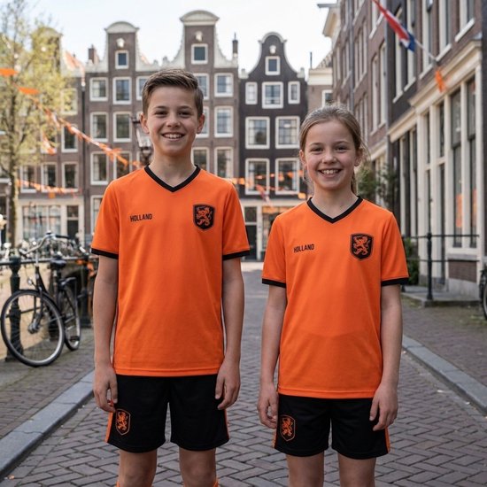 Jongen en meisje in oranje voetbaltenues met 'HOLLAND' opdruk, lachend in een straat met Nederlandse vlaggen.