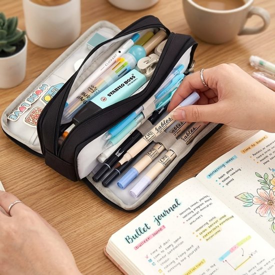 Een vrouw pakt een lichte pen uit een zwart pennenetui, gevuld met pennen en markeerstiften. Naast de etui ligt een bullet journal met bloemenillustraties.