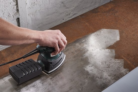 Een hand houdt een groen-zwarte multischuurmachine vast op een metalen plaat.