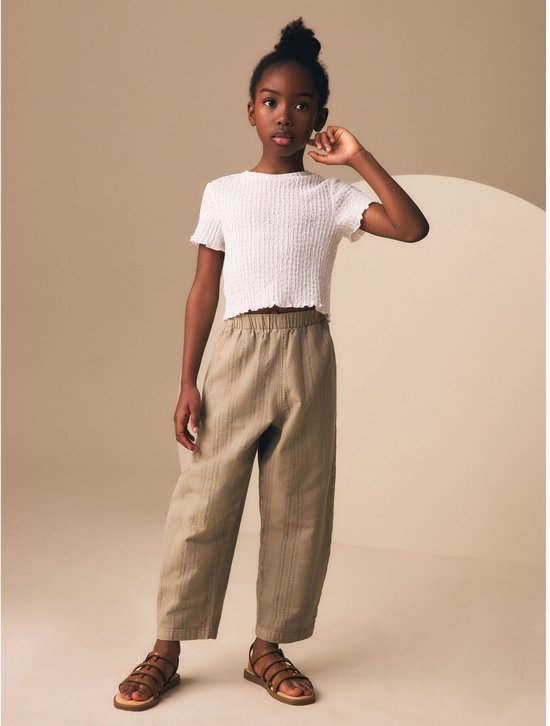 Meisje in een witte top met korte mouwen en een beige broek met verticale strepen, met sandalen.