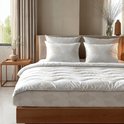 Een opgemaakt bed met een wit dekbed en kussens met beige bladmotieven, bij een raam en houten nachtkastje.