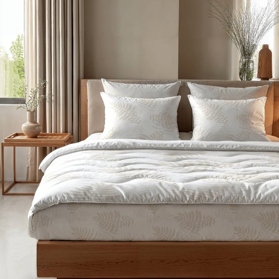 Een opgemaakt bed met een wit dekbed en kussens met beige bladmotieven, bij een raam en houten nachtkastje.