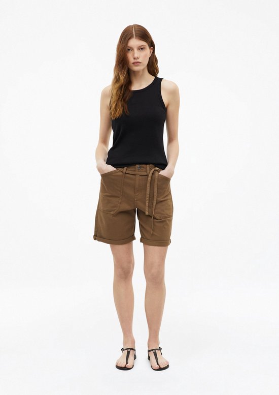 Model in zwarte tanktop en bruine short met riem, handen in de zakken.