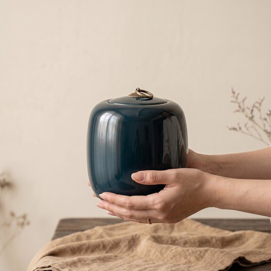 Donkerblauwe, glanzende keramische urn met gouden handvat, vastgehouden in twee handen.