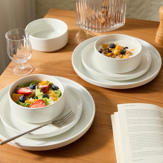Een tafel gedekt met witte borden en kommen, gevuld met fruitsalade en yoghurt met fruit.