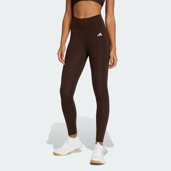 Vrouw in donkerbruine legging met wit Adidas logo, staand, witte sneakers.
