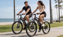 Man en vrouw in sportkleding rijden op zwarte elektrische mountainbikes langs het strand.