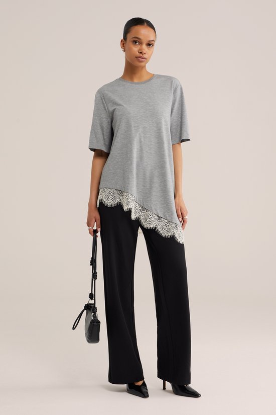Model draagt een zwarte jersey broek met regular fit en een grijs, asymmetrisch shirt met kanten rand.