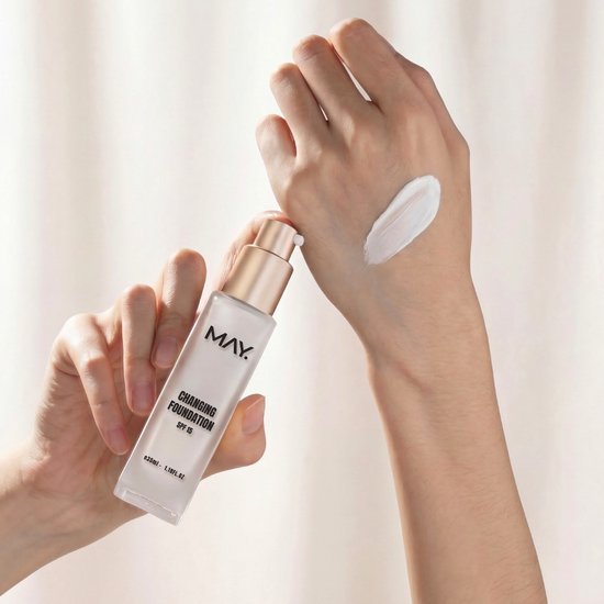 Hand met gouden pompflacon "CHANGING FOUNDATION SPF 15" en een veeg witte foundation op de andere hand.