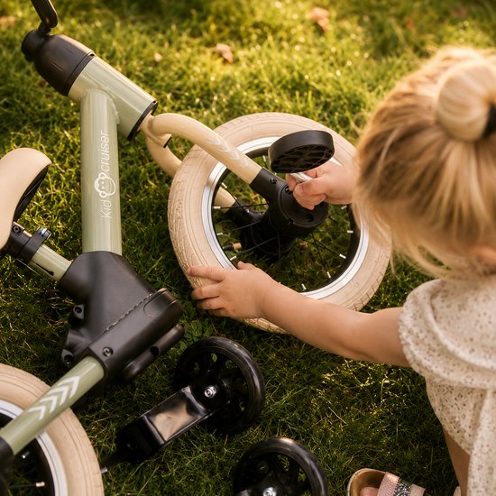 Kind op gras die een lichtgroene loopfiets met creme witte banden aanraakt.